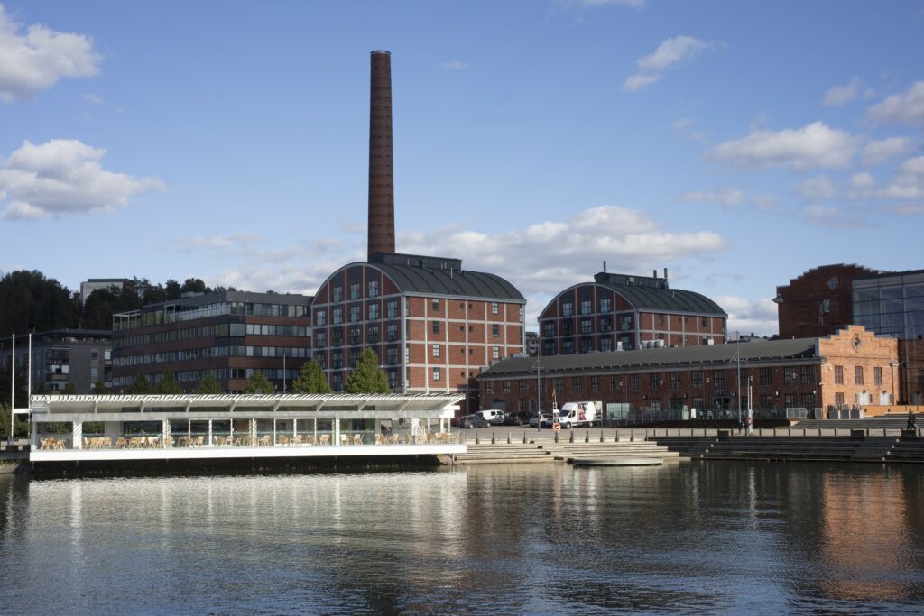 Punatiilisiä rakennuksia rannassa. Rakennusten takana kohoaa piippu, taivas on sininen.